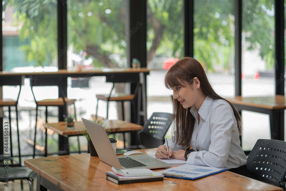Businesswoman writing a note while using laptop at a table in an office. Businesswoman writing a note while using laptop at a table in an office.