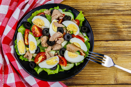 Tasty tuna salad with lettuce, black olives, eggs and fresh vegetables on wooden table. Top view
