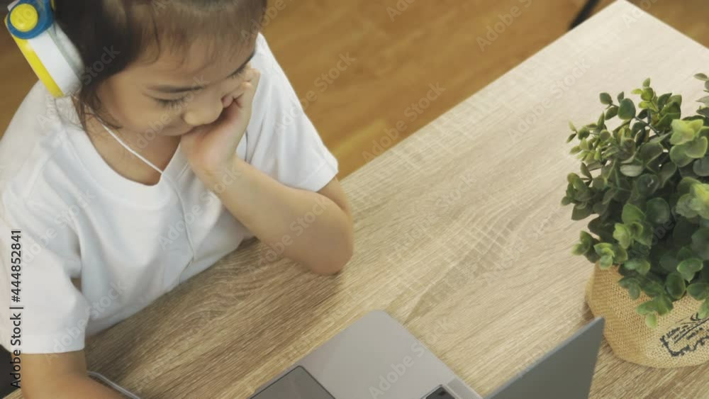 Top view 4K of adorable asian girl kid, wearing headphone with smiling happy face, is learning online from computer laptop on wooden desk shows concept of home education using internet technology. Top view 4K of adorable asian girl kid, wearing headphone with smiling happy face, is learning online from computer laptop on wooden desk shows concept of home education using internet technology.