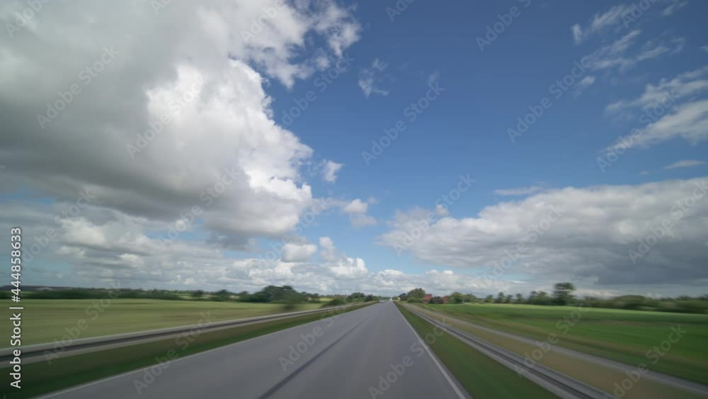 Dashcam view. Driving on the narrow two-lane rural asphalt road with oncoming traffic through the villages and green farm fields. Fluffy white clouds in the blue sky. Dashcam view. Driving on the narrow two-lane rural asphalt road with oncoming traffic through the villages and green farm fields. Fluffy white clouds in the blue sky.