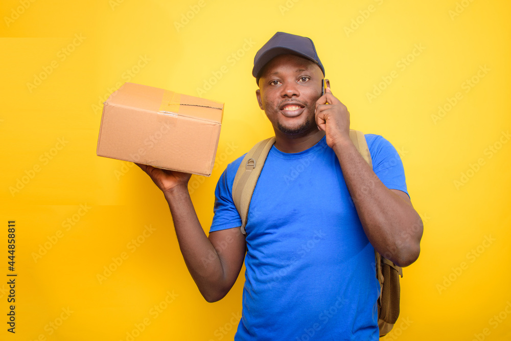 An African dispatch man with back pack, making a call, carrying boxes and wearing a face cap An African dispatch man with back pack, making a call, carrying boxes and wearing a face cap