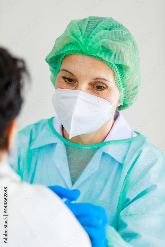 Caucasian senior female doctor in PPE full hazard protection uniform with face mask using syringe needle inject vaccine to woman patient shoulder at hospital vaccinating desk Caucasian senior female doctor in PPE full hazard protection uniform with face mask using syringe needle inject vaccine to woman patient shoulder at hospital vaccinating desk