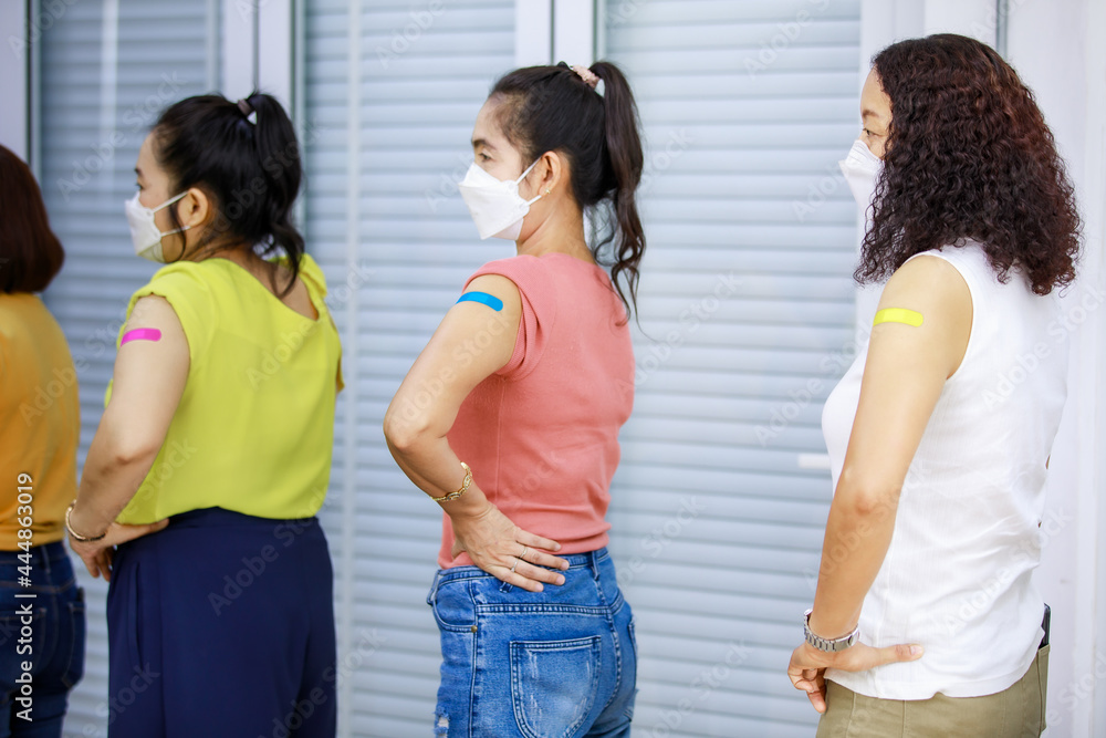 Three Asian female patients standing waiting in orderly straight line showing colorful plasters on shoulder after receiving Coronavirus Covid-19 vaccine injection shot in hospital already