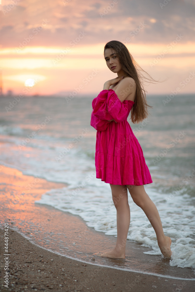 Young girl in a pink summer dress walks on the sea coast at sunrise Young girl in a pink summer dress walks on the sea coast at sunrise