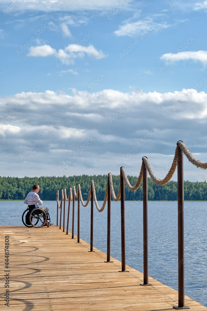 man in wheelchair  fishing on spinning rod on summer day on the pier made of rough boards, background - clouds in the summer sky and a forest lake