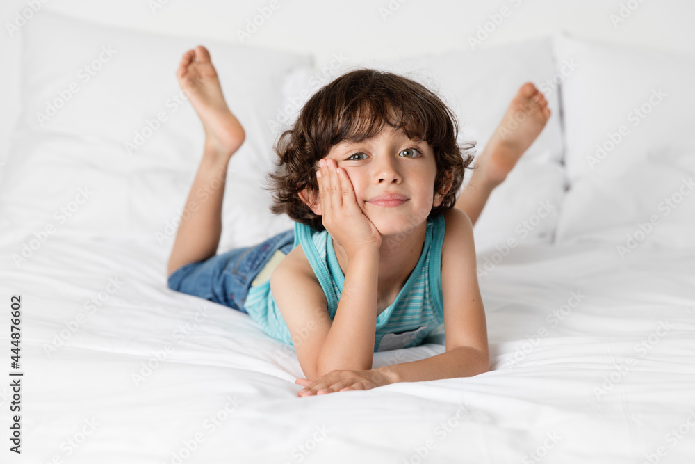 Young boy lying on white bed resting on elbows with face on hands Young boy lying on white bed resting on elbows with face on hands