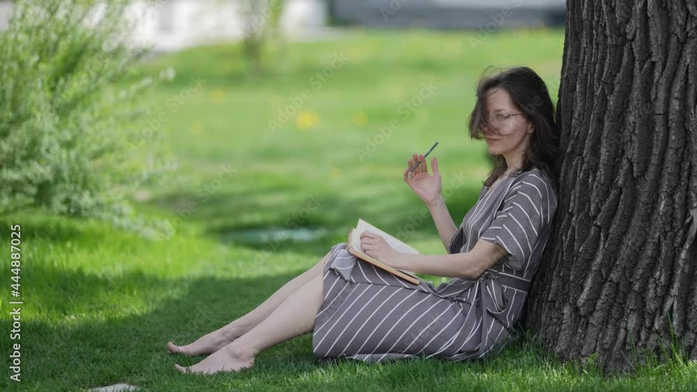 pretty adult woman is sitting near tree with book, resting and dreaming pretty adult woman is sitting near tree with book, resting and dreaming