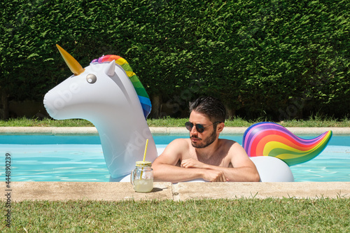 Young man wearing sunglasses into a unicorn inflatable ring in a swimming pool with a refreshment. Summer concept. © Ladanifer