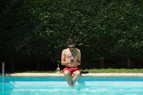 Young man wearing sunglasses using his smartphone sitting on the swimming pool edge. Summer concept.