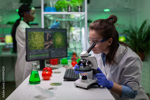 Portrait of biologist scientist in white coat working in expertise laboratory looking into microscope analyzing organic gmo leaf. Specialist researcher doing biochemistry experiment.