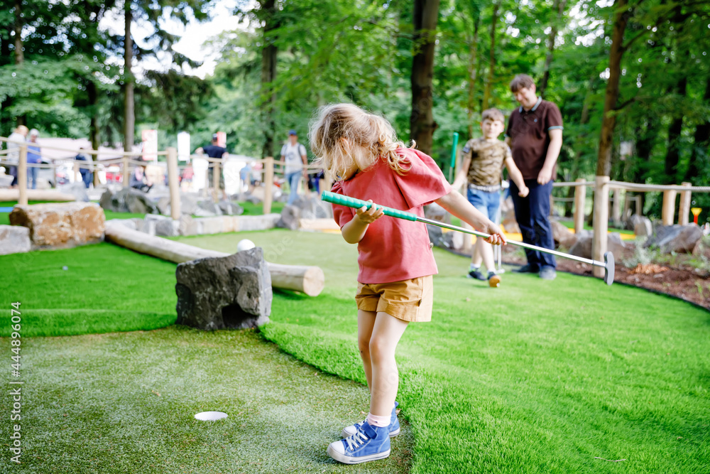 Cute preschool girl playing mini golf with family. Happy toddler child having fun with outdoor activity. Summer sport for children and adults, outdoors. Family vacations or resort. Cute preschool girl playing mini golf with family. Happy toddler child having fun with outdoor activity. Summer sport for children and adults, outdoors. Family vacations or resort.