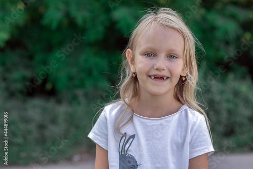 portrait of a girl, a preschooler with blond hair without upper teeth, in the park, a charming child six-year-old blonde on the background of nature laughs cheerfully with a toothless mouth