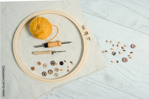Embriodery hoop with two punch needles, ball of yellow yarn, wooden buttons and piece of linnen fabric, with some wooden buttons on white wooden floor. Punch work creative hobby.