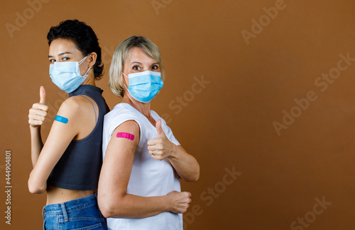 Caucasian female patients wears face mask standing look at camera lean on each other back showing thumb up with colorful plaster together after vaccination in front of brown wall background