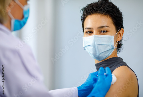 Portrait closeup shot of female patient wears face mask sit looking at camera while doctor wears white lab coat and rubber gloves using syringe needle and cotton injecting coronavirus vaccine