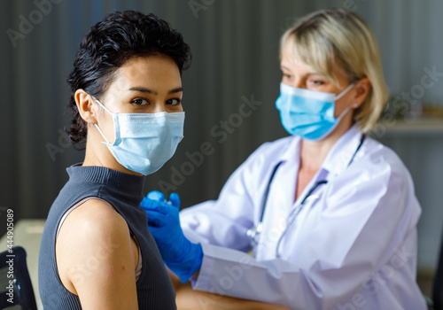 Beautiful short black hair female patient wears face mask sit look at camera while Caucasian doctor in white lab coat with stethoscope injecting vaccine in to her shoulder in blurred background