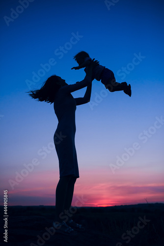 Figure of a woman lifting a child up with the background of evening dark sky and setting sun. Motherhood and parenting.