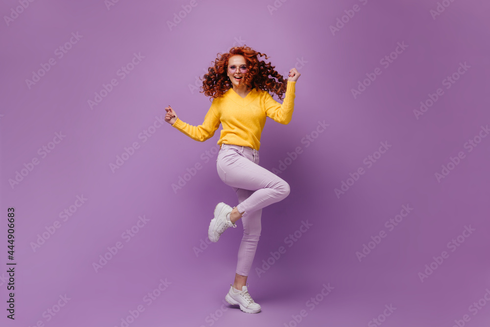 Curly girl in warm sweater and pants jumping and having fun on purple background