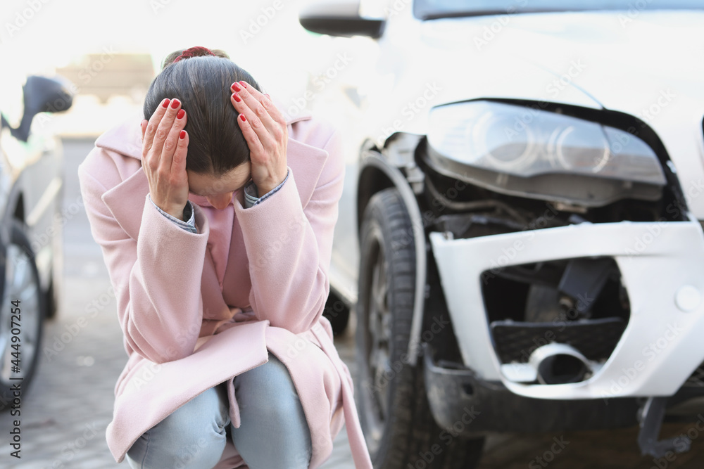 Upset young woman sits with head bowed next to wrecked car