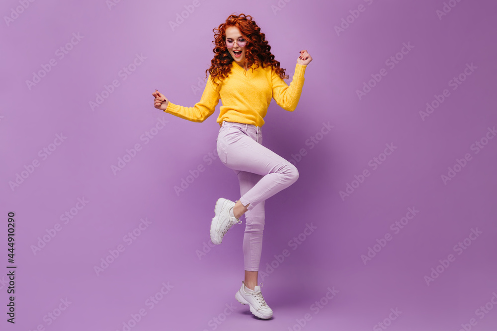 Slender girl in light pants and yellow sweater dances against lilac background