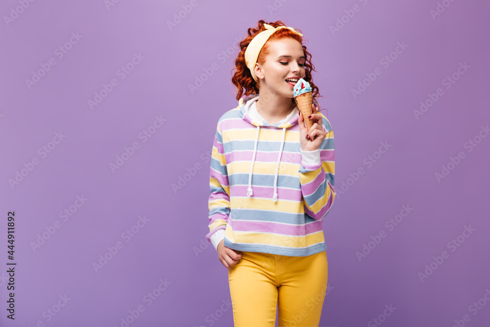 Positive girl in headband and yellow pants licks blue ice cream on isolated background Positive girl in headband and yellow pants licks blue ice cream on isolated background