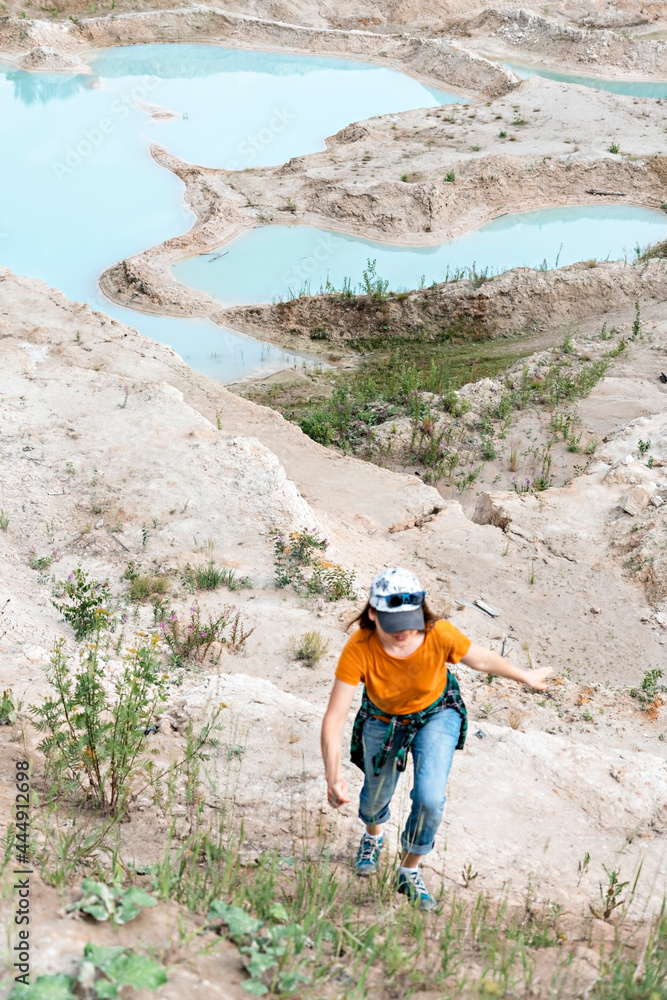 Happy young woman traveler in cap, ginger T-shirt and plaid shirt running on sand of clay quarry with blue turquoise water, beautiful landscape