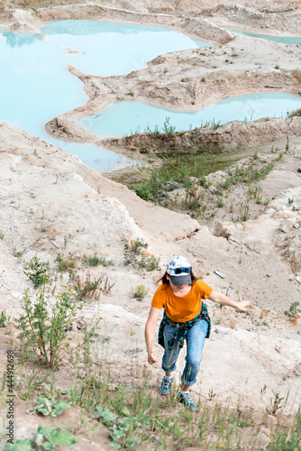 Happy young woman traveler in cap, ginger T-shirt and plaid shirt running on sand of clay quarry with blue turquoise water, beautiful landscape
