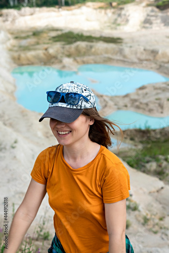 Happy young smiling woman traveler in cap  ginger T-shirt and plaid shirt running on sand of clay quarry with blue turquoise water  beautiful landscape view selective focus