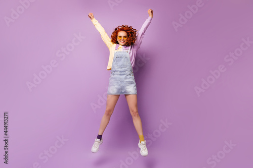 Girl in orange glasses, denim dress and sweatshirt joyfully jumping on purple background