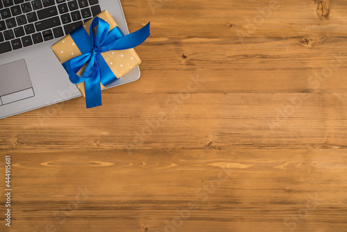 Top view photo of stylish beige small giftbox with blue ribbon bow on laptop on isolated wooden desk background with empty space