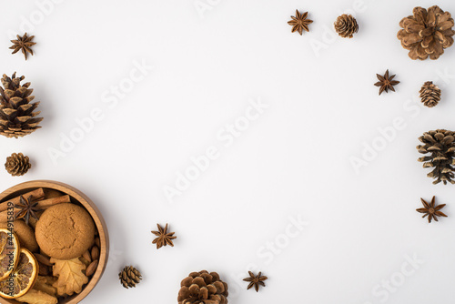 Top view photo of winter composition pine cones anise and wooden bowl with cookies cinnamon sticks and dried lemon slices on isolated white background with empty space in the middle