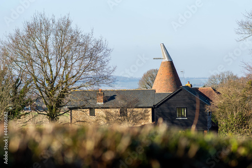 A Kentish oast house with hills (North Downs) visible on the horizon surrounded by trees and framed by a hedge in the foreground. This building would have been used to dry hops used in beer production