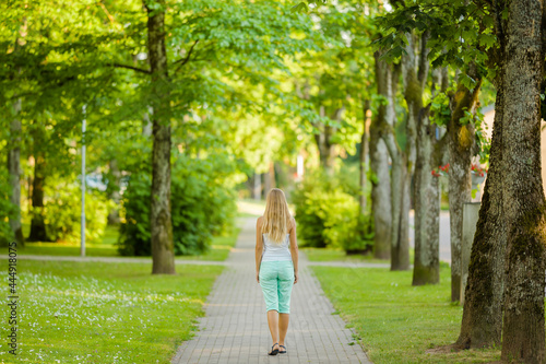 Young adult blonde woman slowly walking on sidewalk at city park in warm sunny summer day. Spending time alone. Peaceful atmosphere. Back view.