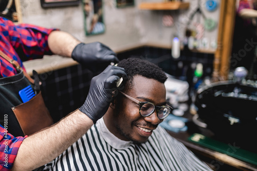 Young African man visiting hairstylist in barber shop. Professional hairdresser cut hair with electric shearer machine. photo
