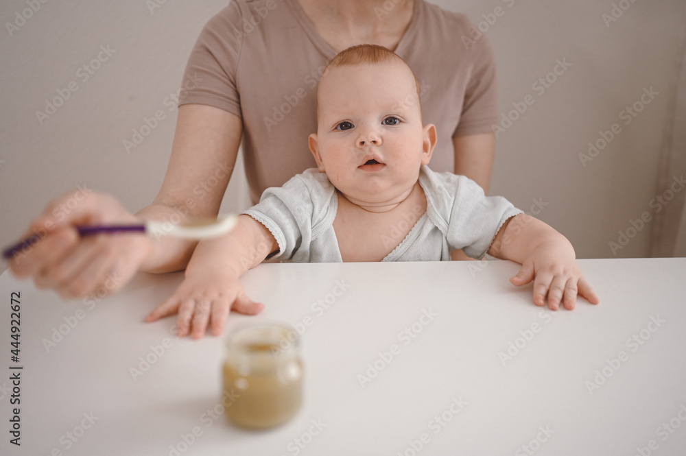 Little newborn funny baby boy learning to eat vegetable or fruit puree from glass jar with spoon. Young mother helping little son eat first food