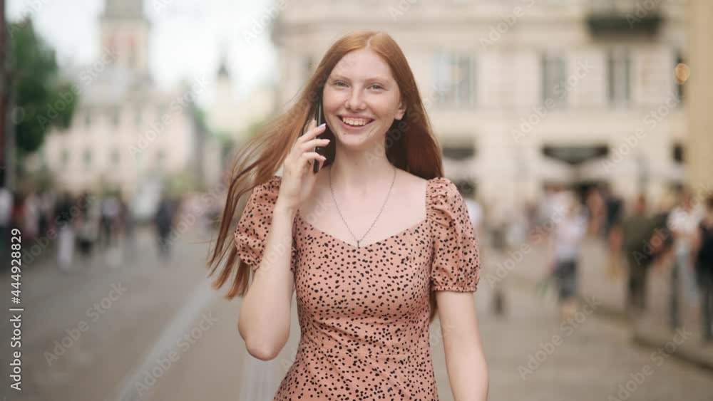 Young beautiful red-haired woman with nice Freckles walking down the city street and talking on the phone. Girl smiling and communicating with friends Young beautiful red-haired woman with nice Freckles walking down the city street and talking on the phone. Girl smiling and communicating with friends