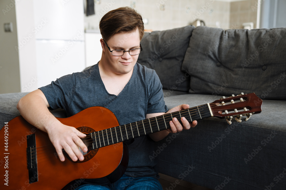 Obraz premium Young man with down syndrome playing guitar while sitting on floor
