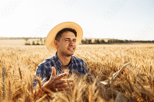 An agronomist checks the field against the backdrop of a summer sunset. Young farmer in a straw hat stands with a tablet and spikelets in his hands in the middle of a ripe wheat field.