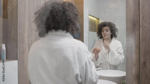 Curly-haired woman applying moisturizer on her face, morning beauty procedures photo