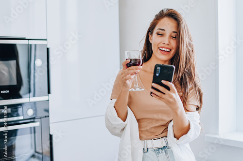 attractive Asian woman drinking wine using a mobile phone and a laptop in the kitchen at home. Writing in a notebook