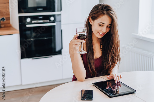 attractive Asian woman drinks wine and using a tablet in the kitchen at home