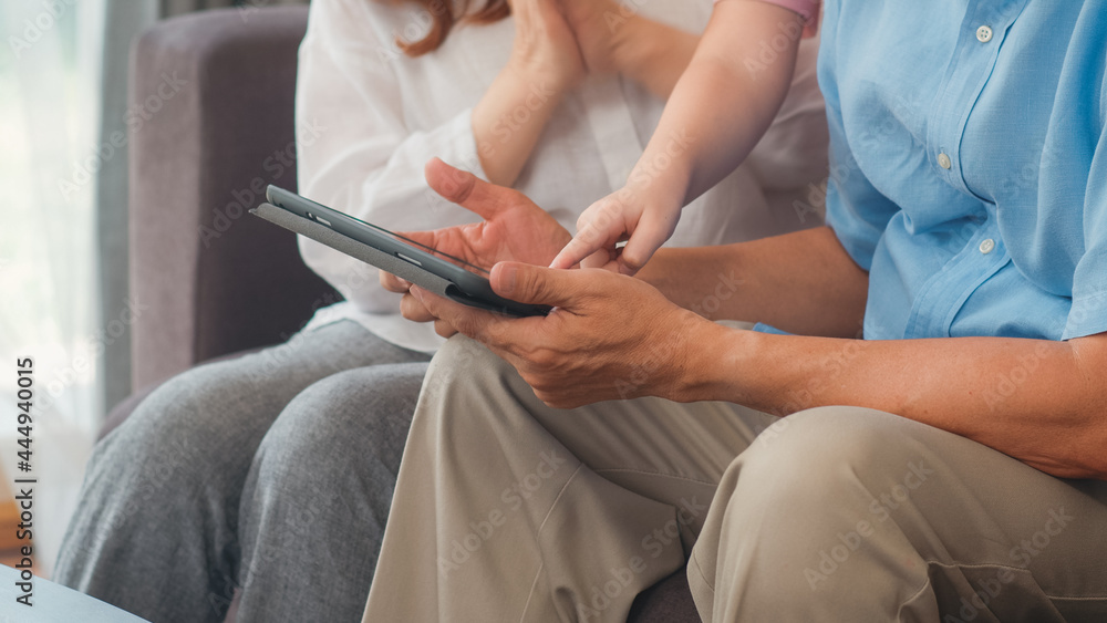 Asian grandparents and granddaughter using tablet at home. Senior Chinese, grandpa and grandma happy spend family time relax with young girl checking social media, lying on sofa in living room concept