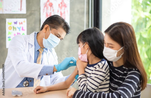 Asian senior doctor wearing gloves and isolation mask is making a COVID-19 vaccination in the shoulder of child patient with her mother at hospital.