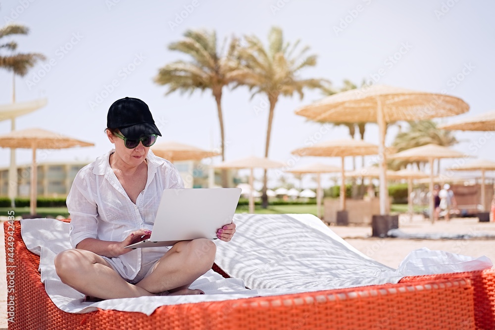 Mature woman using laptop on the background of the beach. Remote work, summer vacation concept, beach work, freelance work and leisure               