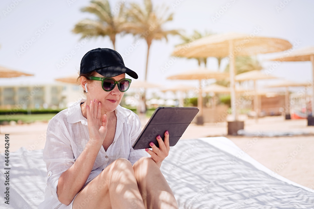 Smiling mature woman having a video chat through tablet on background the beach. Remote work, summer vacation concept, beach work, freelance work and leisure             