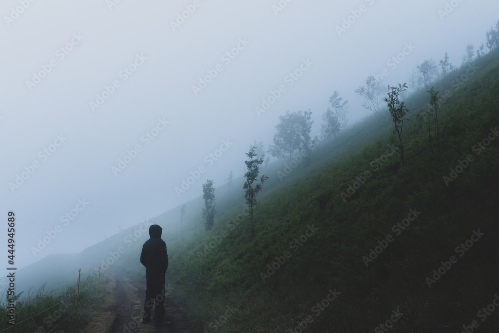 A moody lone hooded figure, back to camera standing on a path Looking at a foggy hillside on a moody atmospheric day.