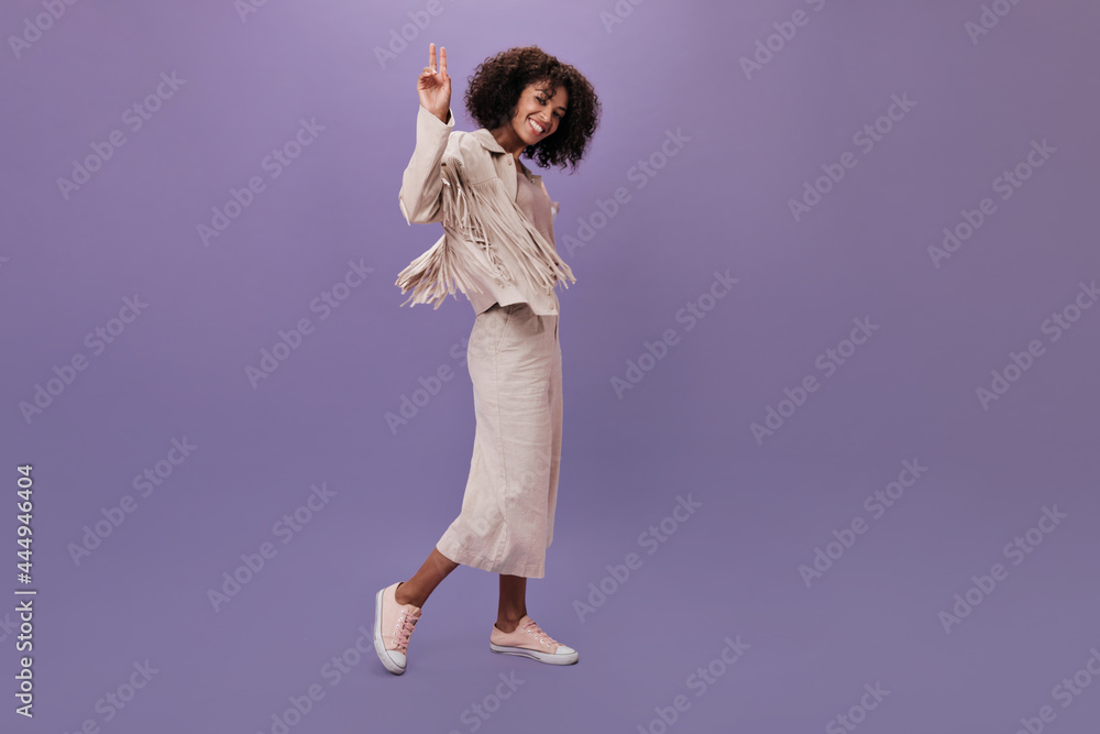 Woman in clothes in style of boho shows sign of peace. Pretty lady in beige jacket and pants dancing and smiling on purple backdrop