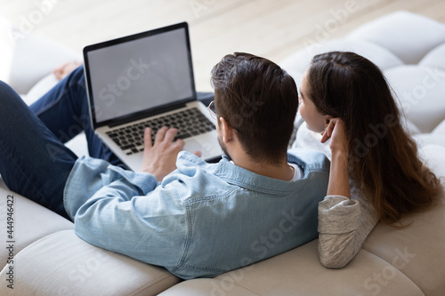 Young spouses sit on sofa relax together at home using computer, buying through e-commerce retail e services, watch new movie online, websurfing, white mock up laptop screen view over couple rear view