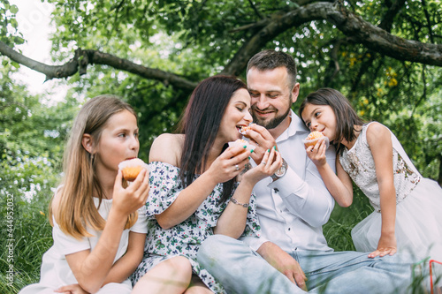 Positive parents with their little daughters sitting on green grass and tasting cupcakes. Happy family enjoying summer picnic on fresh air.