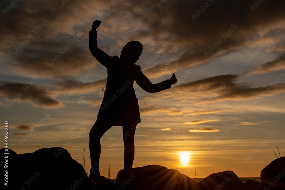 Fototapeta premium Silhouette of a young girl with a triumphantly thrown up hand against the background of the evening sky. World perception concept.